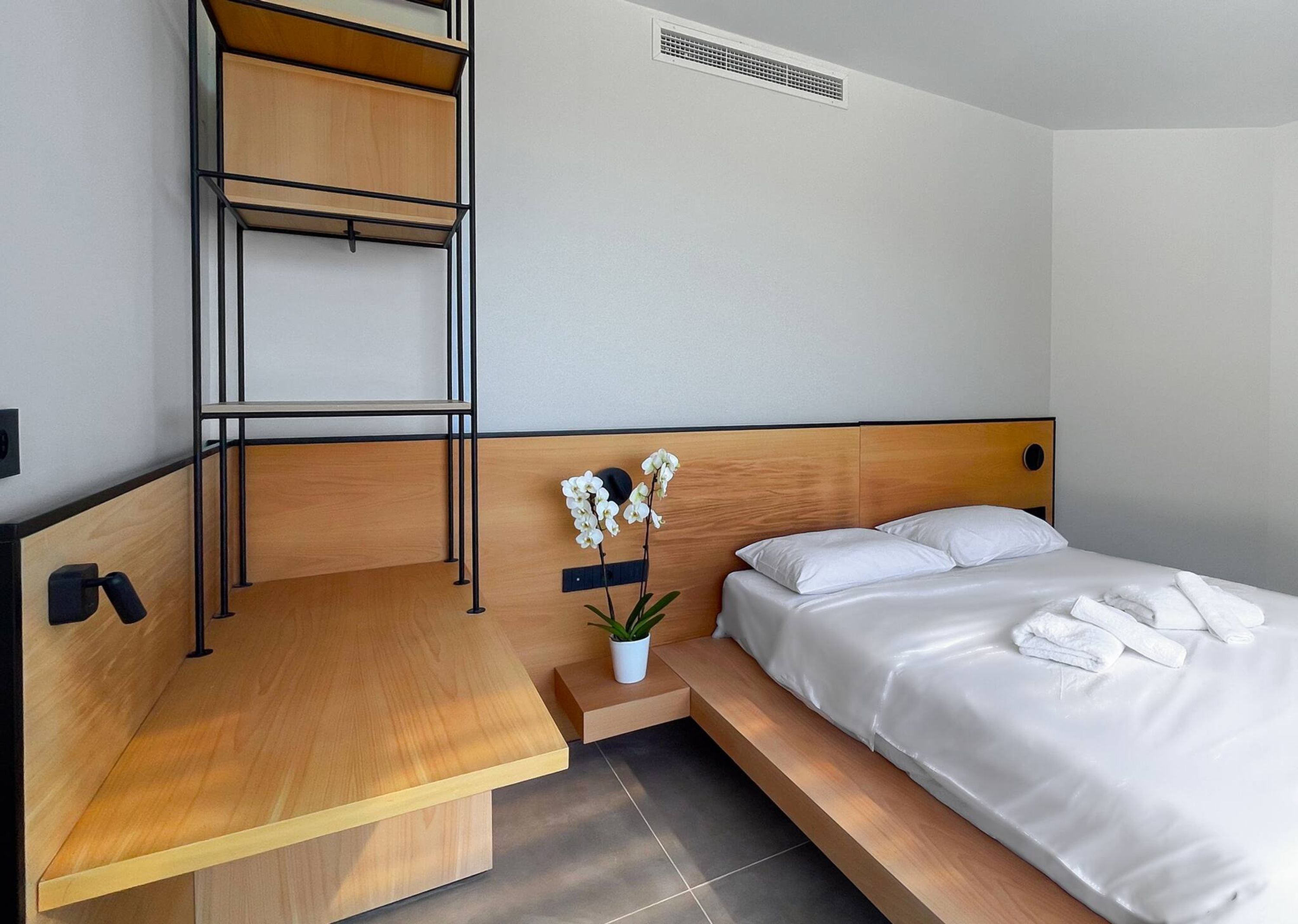 A hotel room with a bed, a desk, a shelf, a potted plant, and a vent.