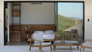 A modern bedroom with a bed, a seating area with chairs and a table, a potted plant, and a view of a landscape through a large window. - Scirocco Telemachos 1 BED (Lefkada)