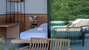 A modern hotel room with a bed, a wooden bench, a small table, and a view of a green landscape through a glass wall. - Scirocco Telemachos 1 BED (Lefkada)