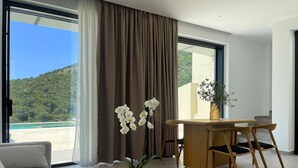 A modern living room with a sofa, a dining table with chairs, a potted plant, and a view of a beach and forest. - Scirocco Telemachos 1 BED (Lefkada)