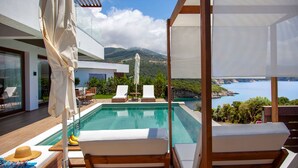 A modern outdoor pool area with lounge chairs, a wooden deck, and a view of the sea and mountains. - Zavia Villa ENA 2B Syvota (Igoumenitsa)