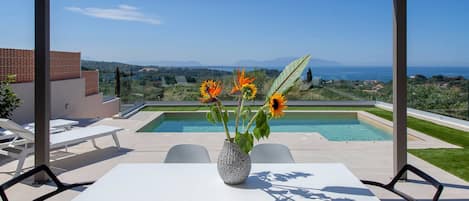 A poolside area with a table and chairs, a vase with flowers, and a view of the sea and hills.