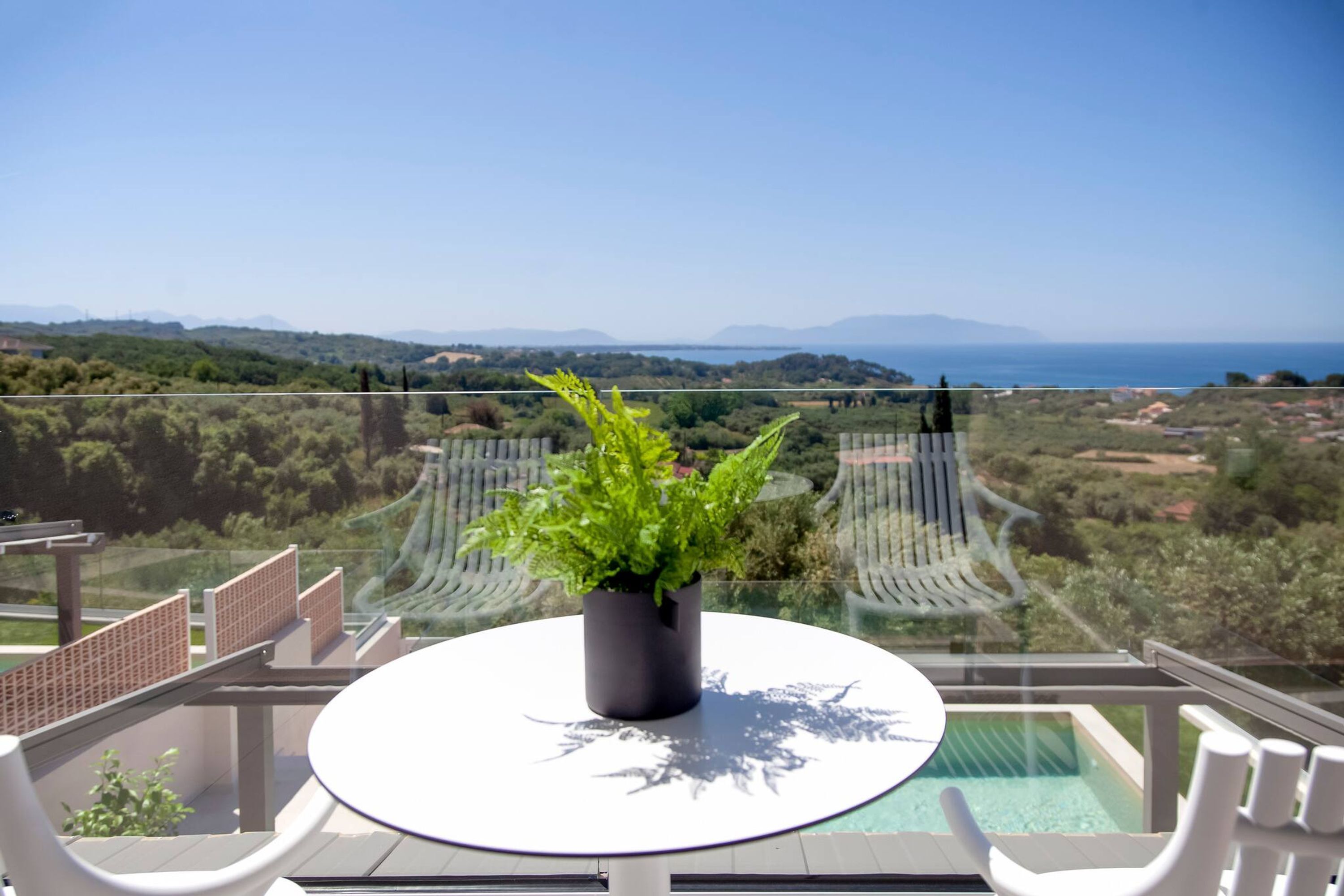 A balcony with a round table, a potted plant, and a clear view of the sea and landscape.