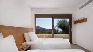 A hotel room with two beds, a wooden headboard, a nightstand with a book and glasses, and a large sliding glass door leading to a balcony with a view of the sea and mountains. - Villa Eptanisa in Sivota (Lefkada)