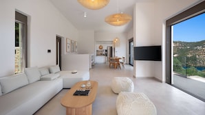 A modern living room with a sofa, coffee table, and ottoman, a dining area, and a large window with a view. - Villa Eptanisa in Sivota (Lefkada)