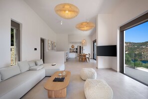 A modern living room with a sofa, coffee table, and ottoman, a dining area, and a large window with a view. - Villa Eptanisa in Sivota (Lefkada)