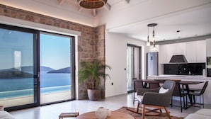 A modern living room with a large sliding glass door to a balcony, a dining area with a table and chairs, and a kitchen with white cabinets. - Smirties Villa Pasithea 4bed (Lefkada)