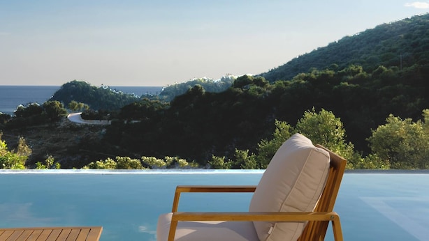 A wooden chair with a cushion by an infinity pool overlooking a coastal landscape with a winding road and dense greenery. - Scirocco Odyssey 2 BED (Lefkada)