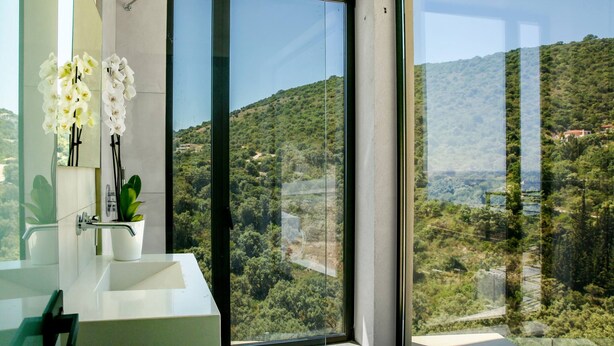 A modern bathroom with a large window, a white sink, and a potted plant. - Scirocco Odyssey 2 BED (Lefkada)