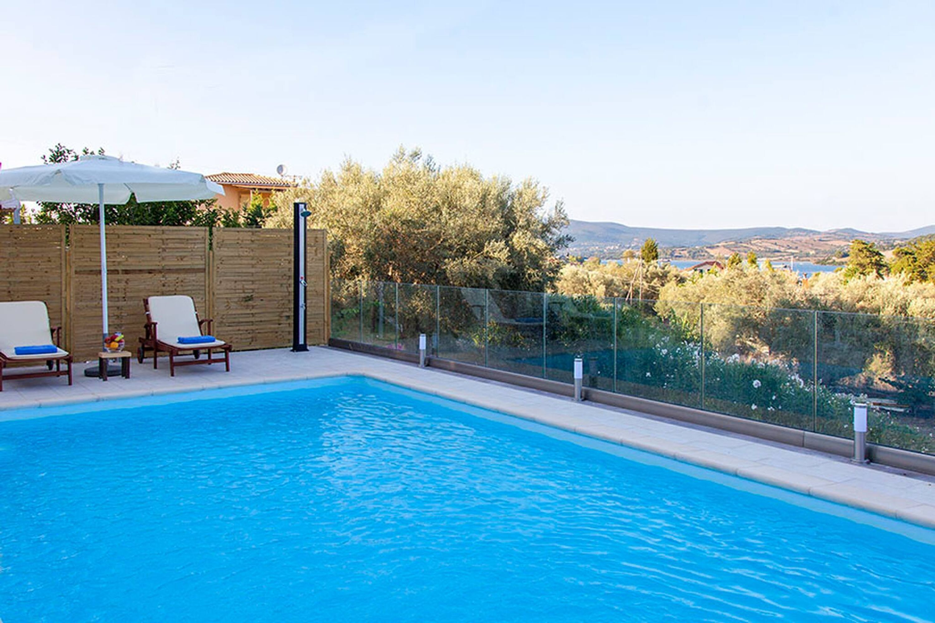 A rectangular swimming pool with clear blue water, surrounded by a wooden fence and a glass barrier. There are lounge chairs with blue cushions and a white umbrella.