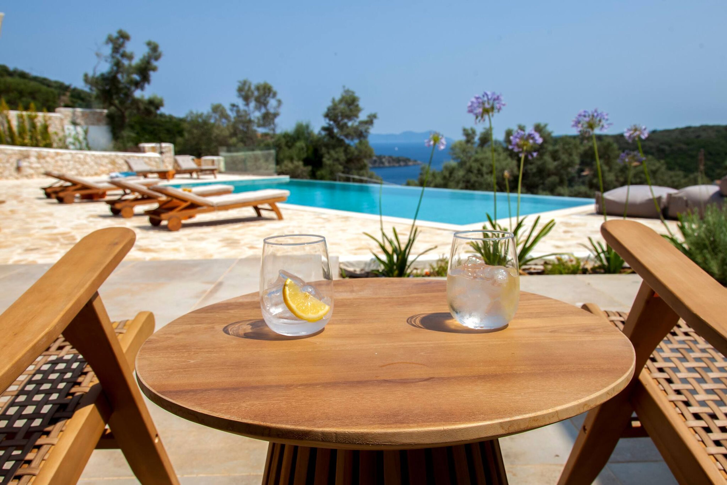 Une table en bois avec deux verres d’eau et une tranche de citron, donnant sur une piscine et offrant une vue panoramique sur des collines et des arbres.