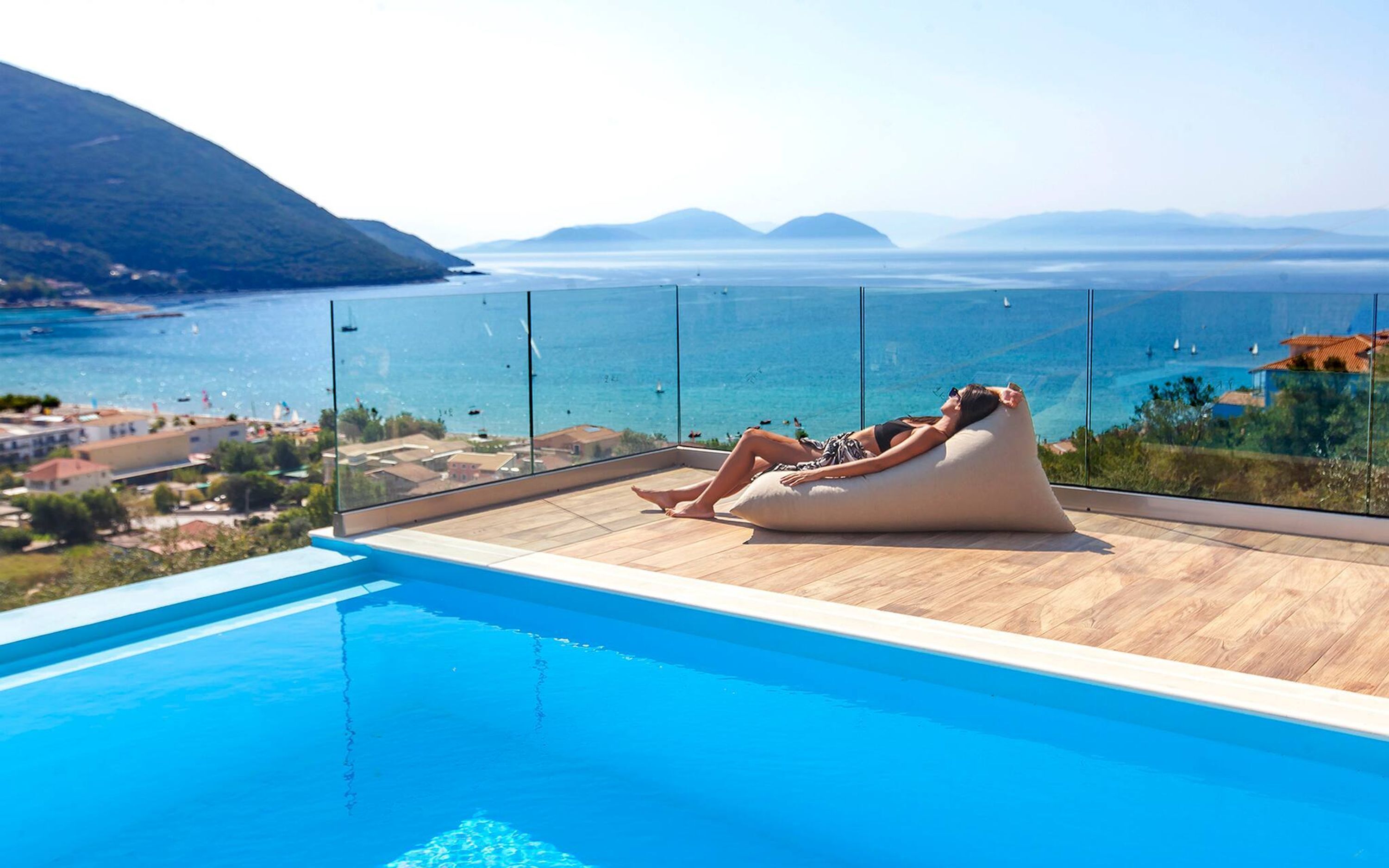 Une personne se détend sur un fauteuil en forme de sac de haricots au bord d’une piscine, avec vue sur la mer et les montagnes.