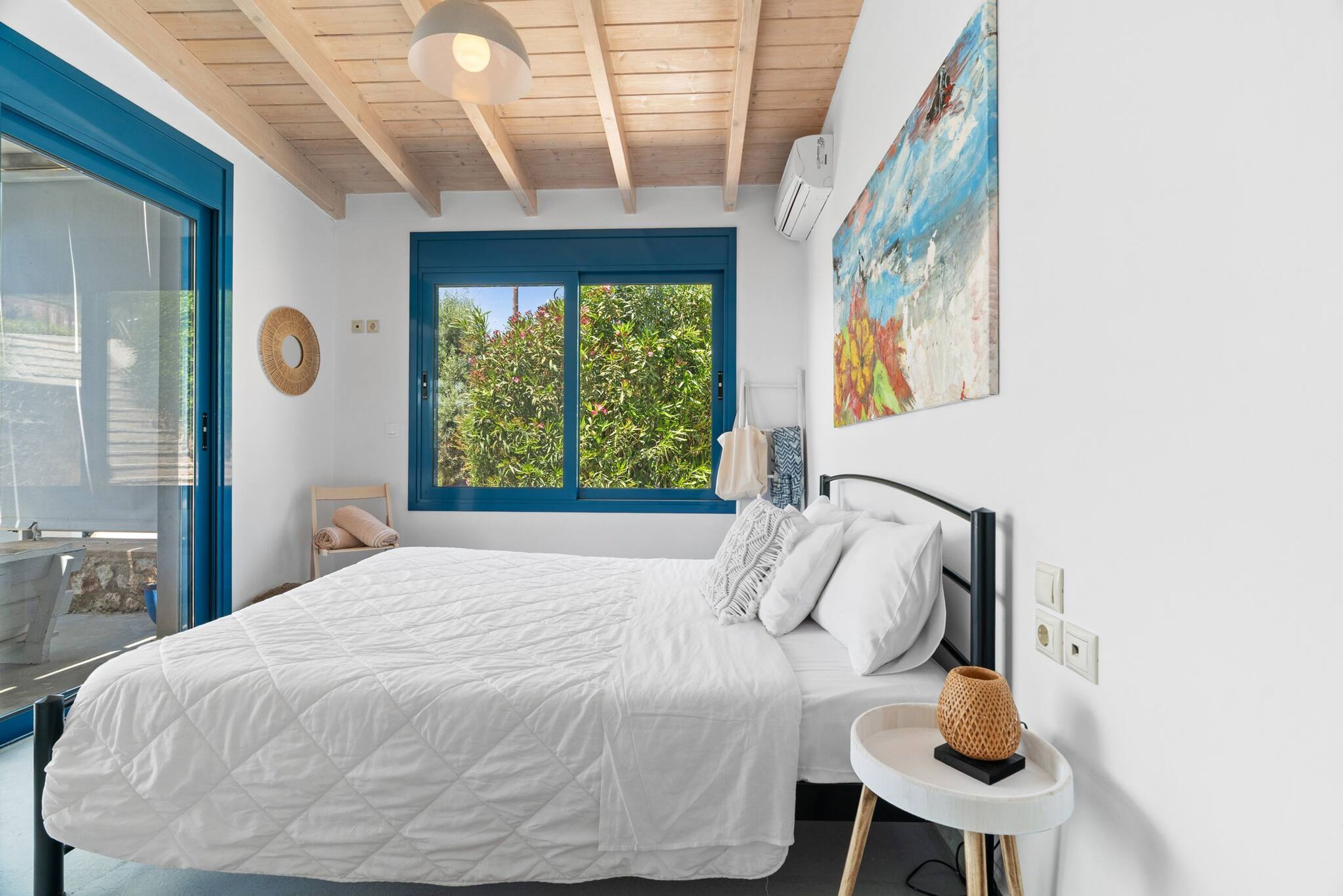 A bedroom with a bed, a window with a view of greenery, a painting on the wall, and a small bedside table.