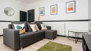 A modern living room with a grey sectional sofa, a round mirror, framed artwork, a small table, and a radiator. - 3 Mins to Stadium W M Dishwasher Sofa Bed (Liverpool)