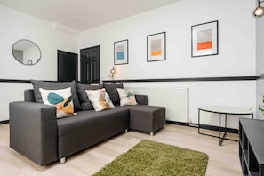 Uma sala de estar moderna com um sofá modular cinza, um espelho redondo, obras de arte emolduradas, uma mesinha e um radiador.