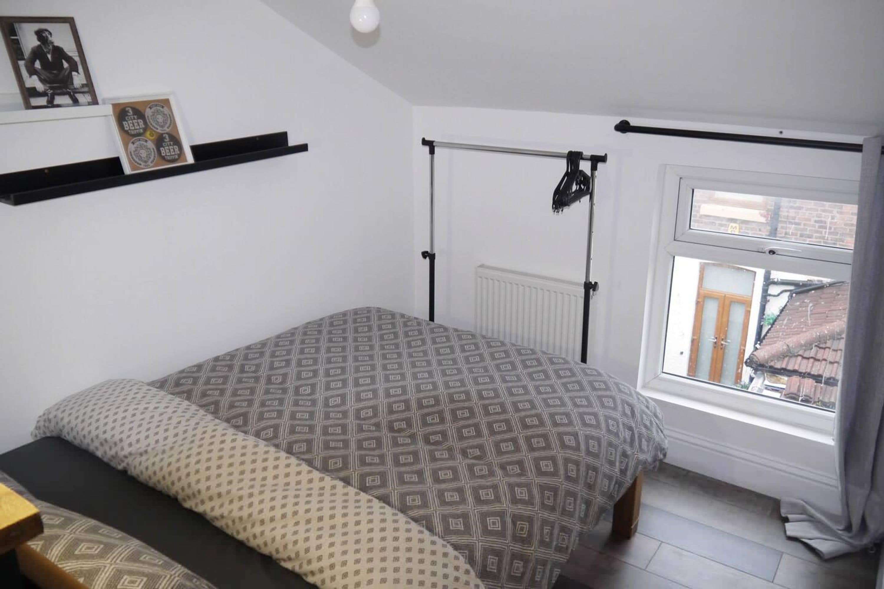 A bedroom with a bed, a radiator, a window with curtains, a shelf with items, and a picture on the wall.