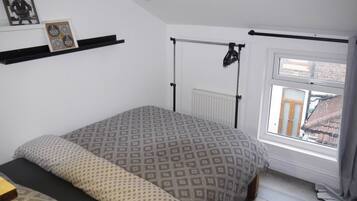 A bedroom with a bed, a radiator, a window with curtains, a shelf with items, and a picture on the wall.