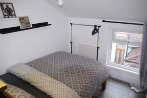 Un dormitorio con una cama, un radiador, una ventana con cortinas, una estantería con objetos y un cuadro en la pared.