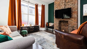 A living room with a brick fireplace, a sofa, a television mounted on the wall, and a window with curtains. - 3 Floors Loft 2mins to Train Patio Parking (Liverpool)