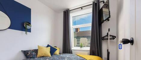 A bedroom with a bed, pillows, a hanging plant, a window with curtains, and a view of the outside.