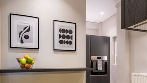 Cozinha moderna com bancada preta, forno de aço inoxidável e geladeira preta. Há duas obras de arte emolduradas na parede e um banquinho com uma tigela de frutas.