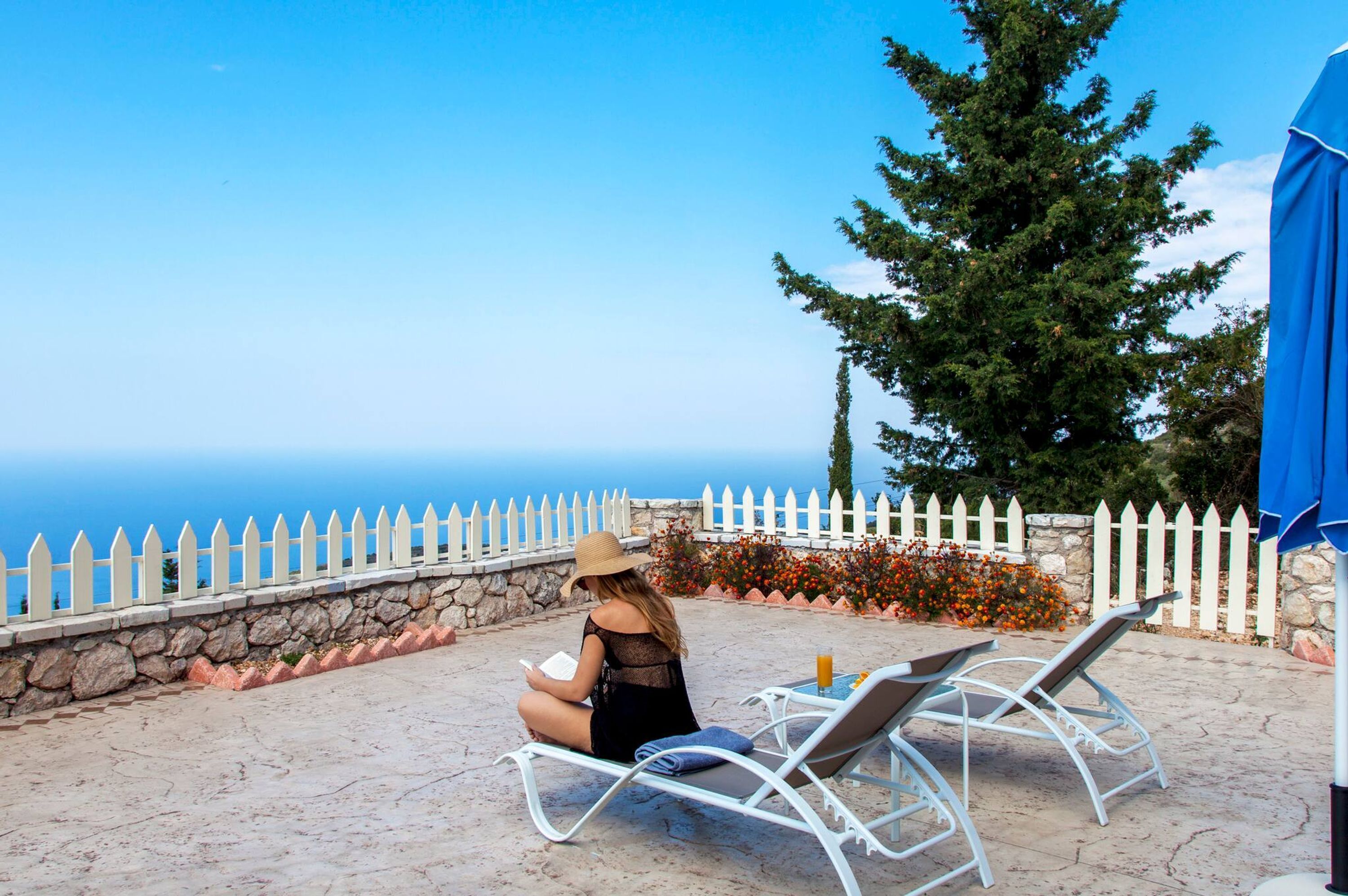 Une femme est assise sur une terrasse, en train de lire, avec vue sur la mer, une clôture blanche en bois et des meubles d’extérieur.