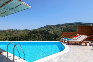A swimming pool with a view of a forested hill, a lounge chair, and a wooden fence. - Villa Alba in Lefkada (Lefkada)