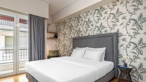 A bedroom with a large bed, a grey headboard, a window with curtains, a small side table, and a patterned wallpaper. - Beehive Ground in Athens (Athens)