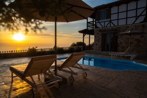 Zona de piscina con sillones reclinables y sombrillas, un edificio de piedra con balcón y vistas al atardecer sobre el agua.