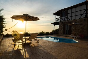 Un área junto a la piscina con sillones y una sombrilla, un edificio de piedra y una vista despejada del atardecer.