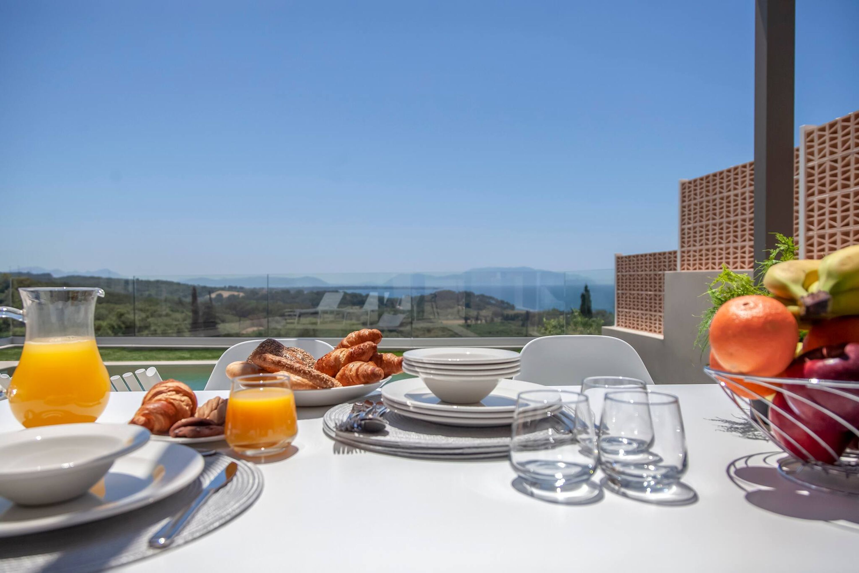 A breakfast table with croissants, orange juice, and fruit, overlooking a scenic landscape.
