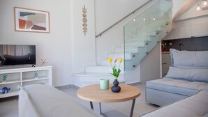 Ett modernt vardagsrum med en platt-TV, ett runt soffbord i trä, en soffa, en trappa med glasräcken och en minimalistisk inredning.