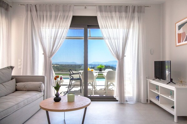 Un salon moderne avec un canapé, une table ronde ornée d’un vase, un téléviseur posé sur un meuble et un balcon avec des chaises et une table.