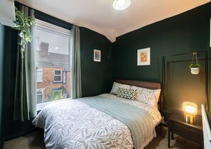 A bedroom with a bed, a window, a bedside table, and a wall-mounted light fixture. - Work-friendly Sefton Penny Lane Free Parking (Liverpool)