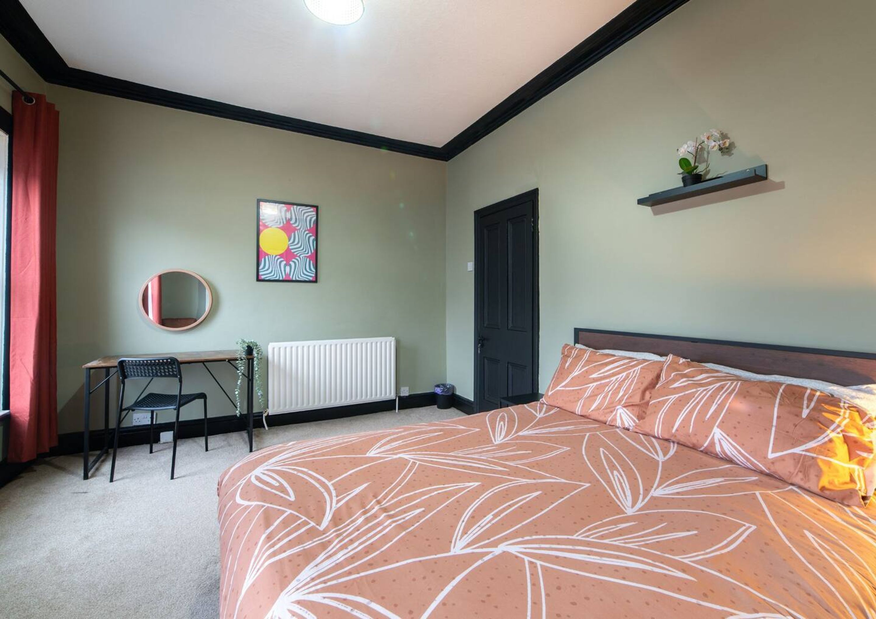 A bedroom with a bed, a desk, a chair, a radiator, a door, and a wall-mounted shelf with flowers.