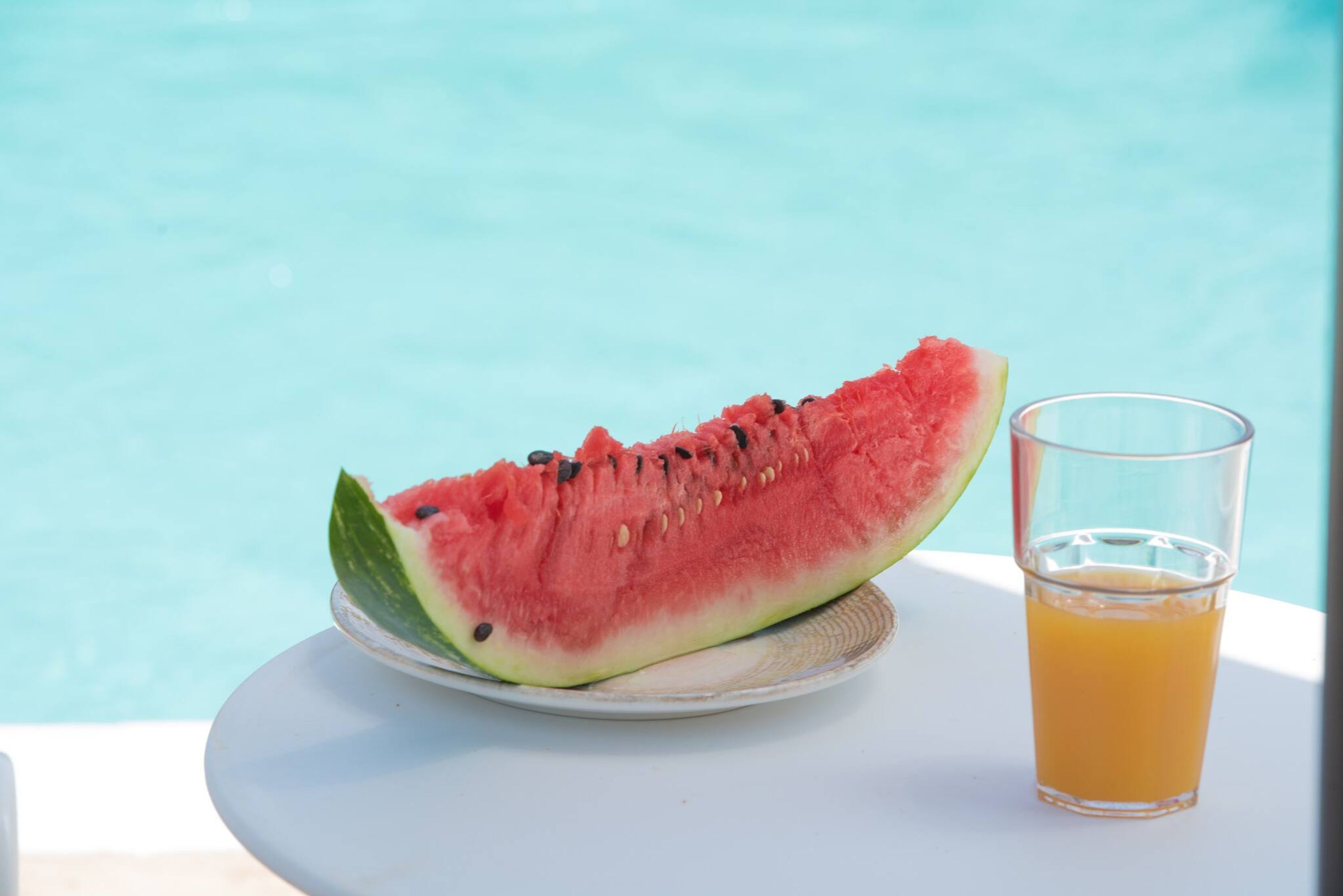 A slice of watermelon and a glass of orange juice on a white table by a pool.