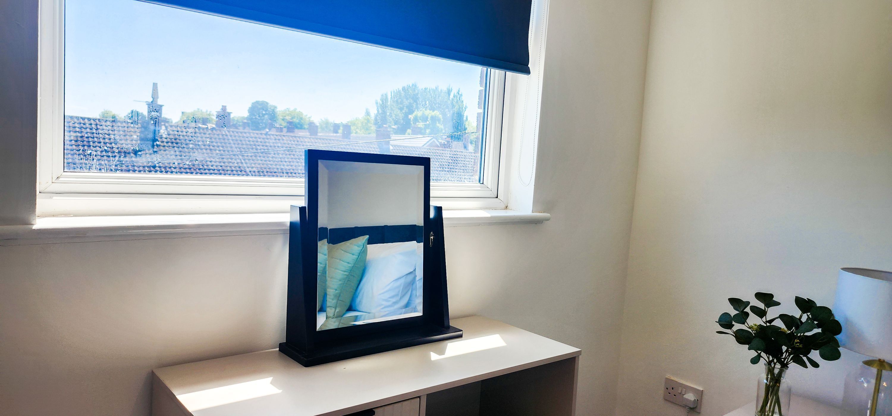 Een kamer met een raam, nachtkastje, bedlamp en een vaas met bloemen.
