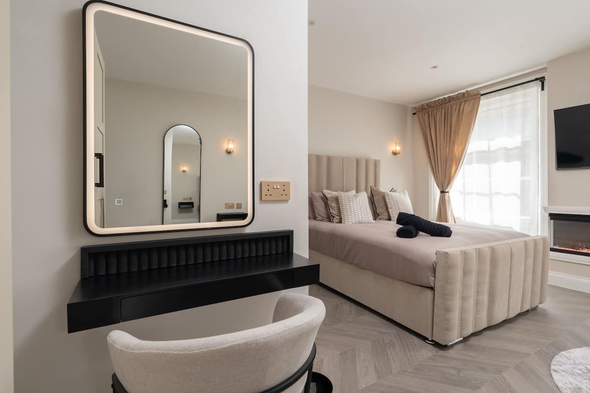 Una habitación de hotel moderna con un espejo grande, una cama con ropa de cama color beige, una consola negra, una silla y una chimenea.