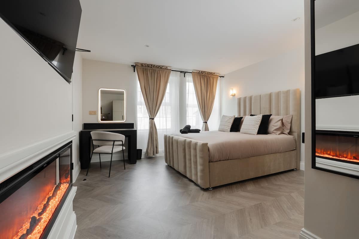 Un dormitorio moderno con cama, escritorio, sillón, chimenea y un amplio ventanal con cortinas.