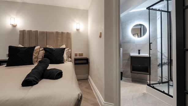 Quarto de hotel moderno com cama, mesas de cabeceira, um espelho circular na parede e porta de vidro que dá para o banheiro.