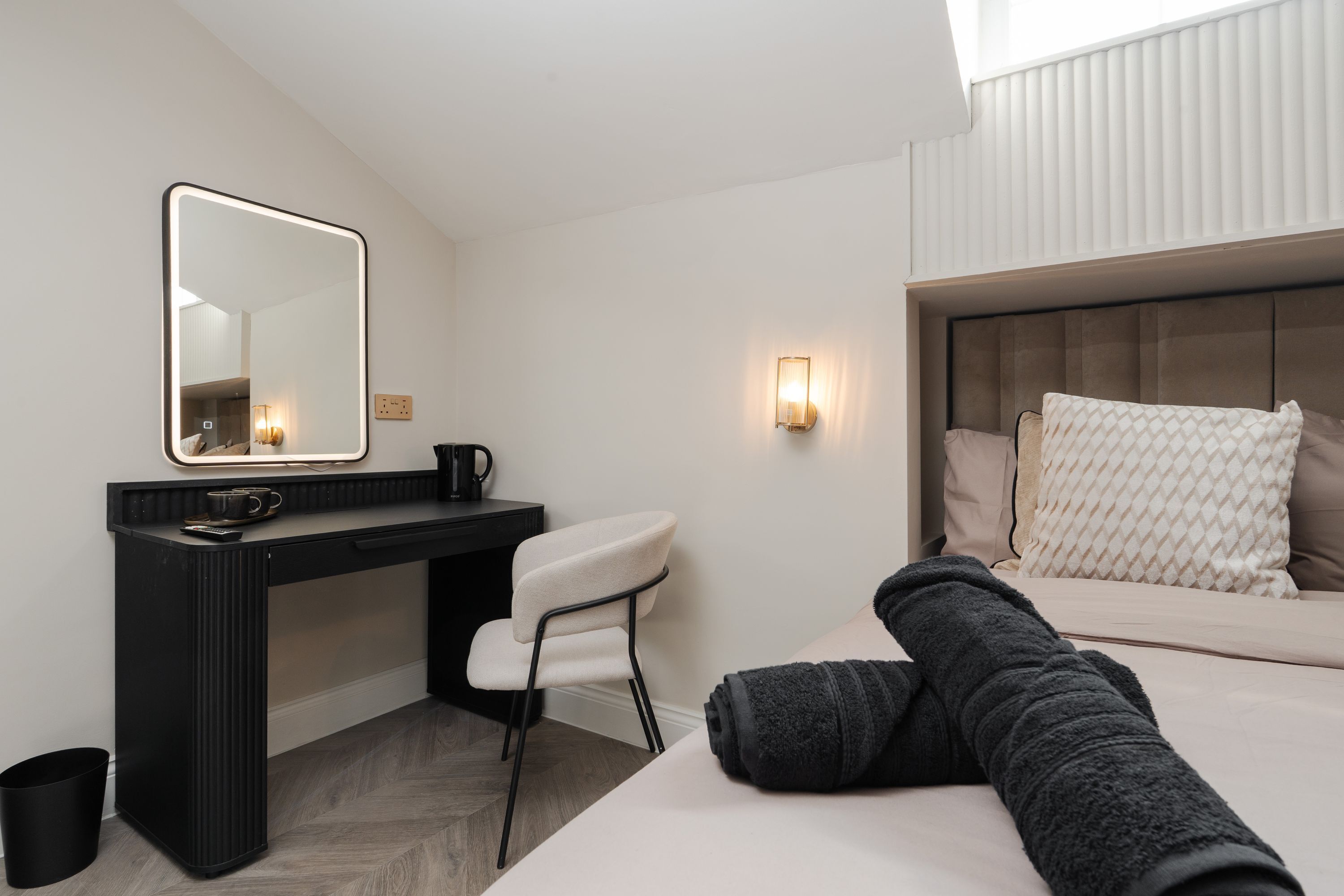A hotel room with a bed, a desk with a chair, a mirror, and a wall-mounted light.