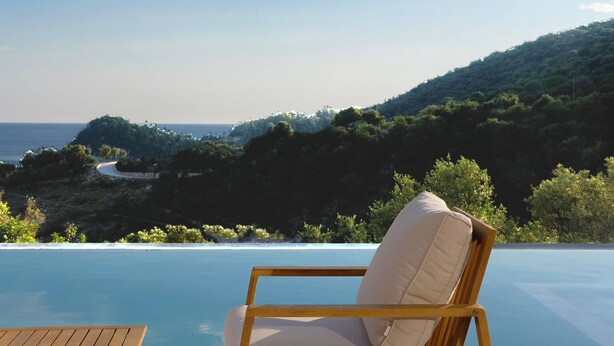 A wooden chair with a cushion by an infinity pool overlooking a coastal landscape with a winding road and dense greenery. - Scirocco Penelope 2 BED (Lefkada)