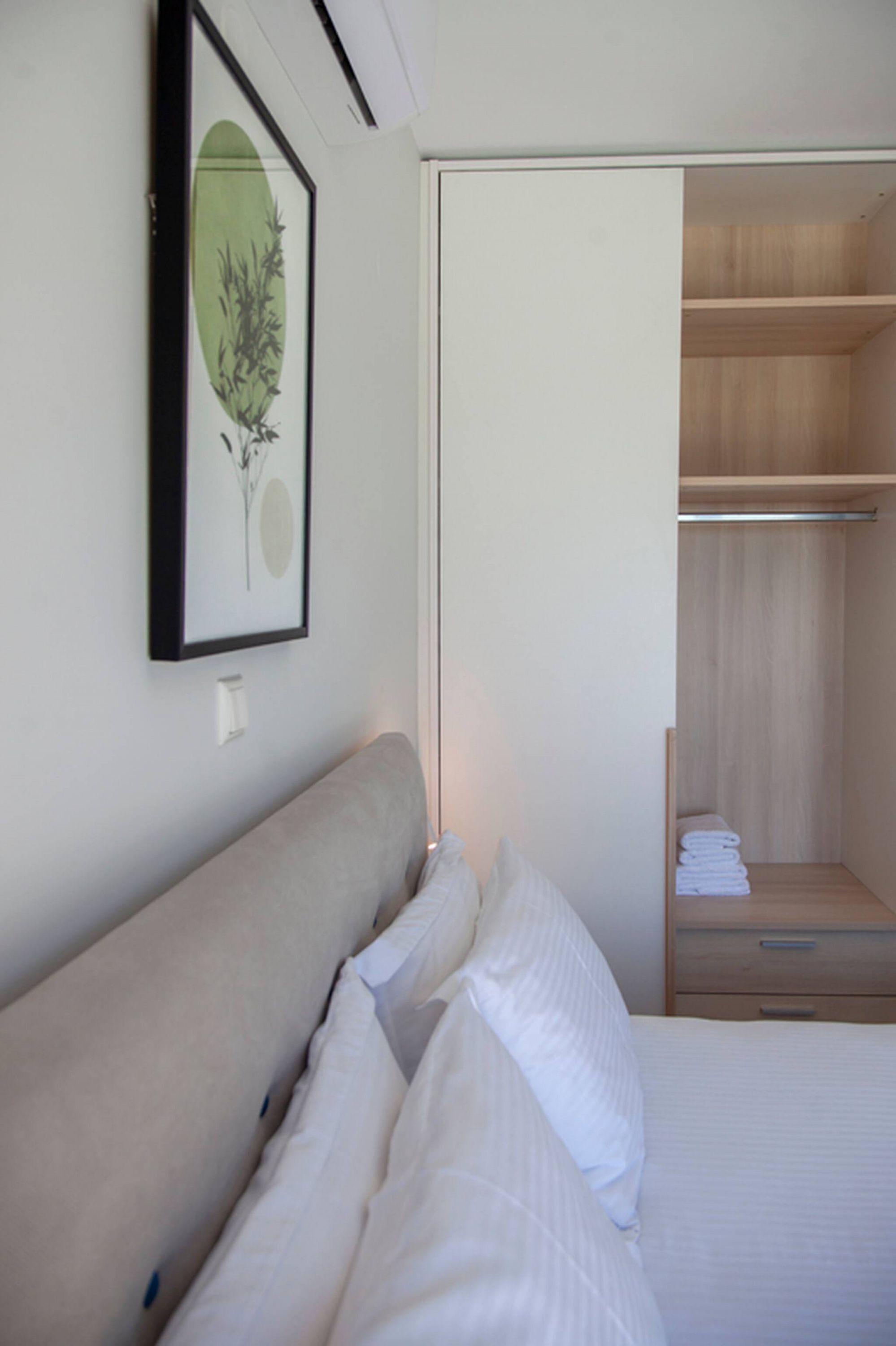 A neatly made bed with white linens, a framed artwork on the wall, and a wooden closet with folded towels.