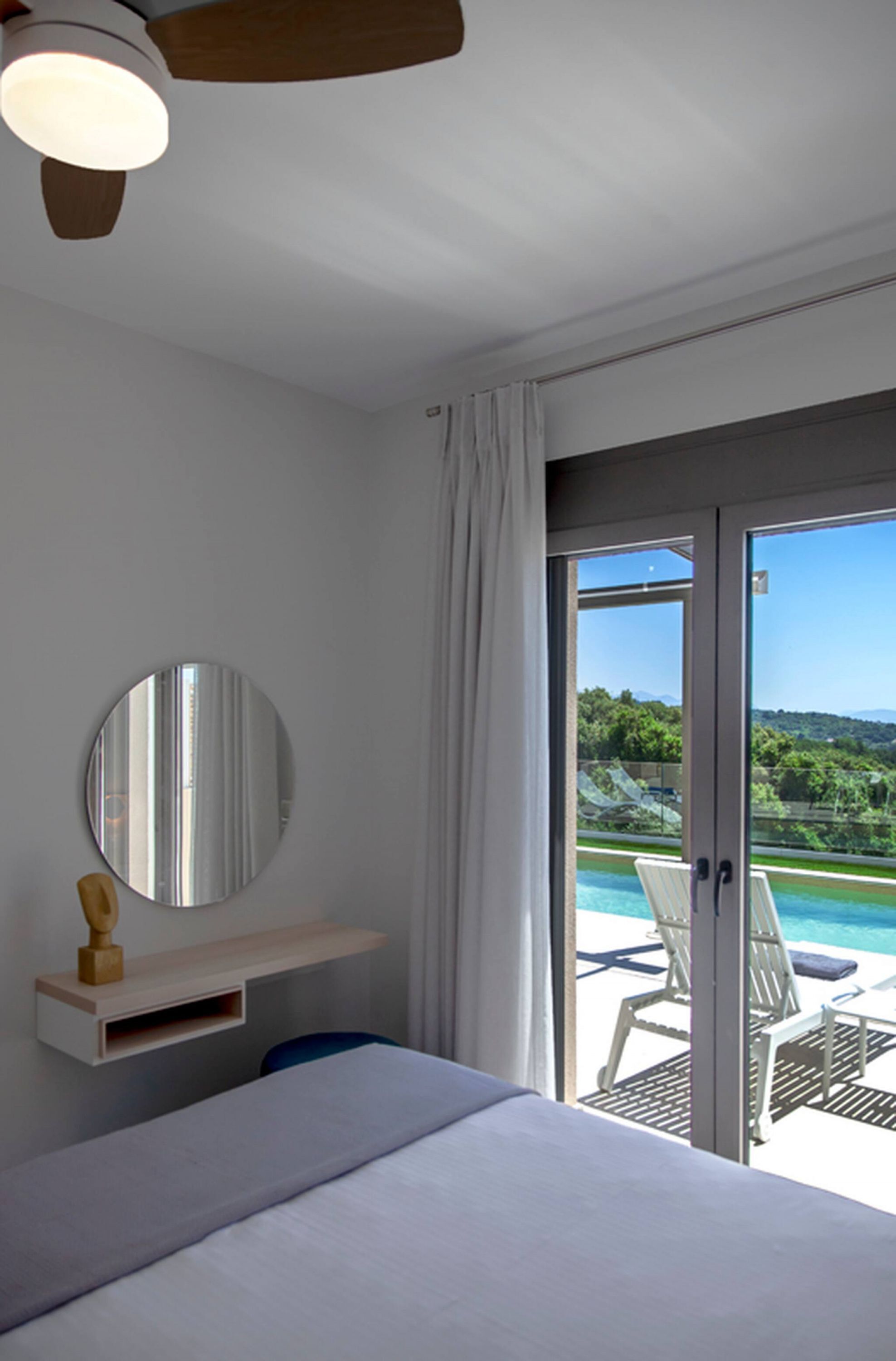 Una camera da letto con un letto, un comodino con uno specchio, una ventola a soffitto e una porta scorrevole in vetro che dà su un balcone con piscina e sedie.