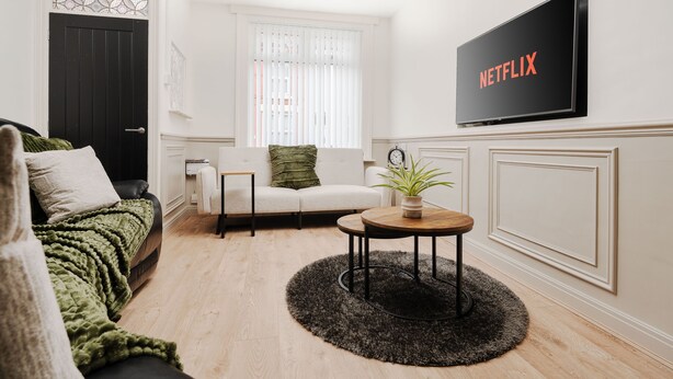 A living room with a black sofa, a white couch, a round coffee table, a TV on the wall, and a window with blinds. - Anfield 3 Mins Superfast Wifi Washer Sofabed (Liverpool)