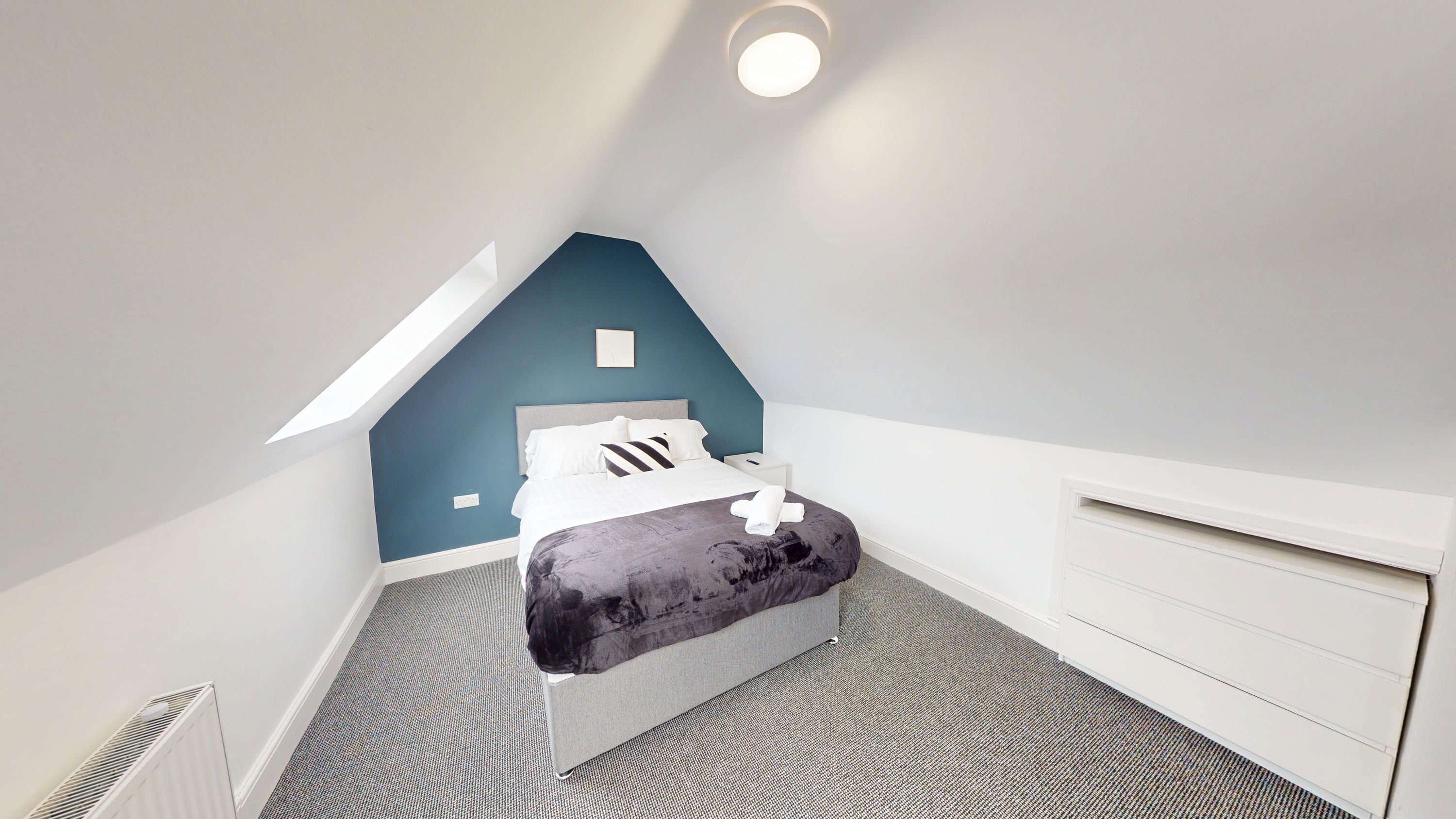 Une chambre avec un plafond en pente, un lit, une table de chevet, une commode et un radiateur.