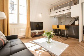 A modern living room with a sofa, a coffee table with a vase, a TV, and a kitchen area with a counter and stools. - Historic Parisian Gem Chic Central Retreat (Paris)