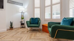 Ein Zimmer mit einem Fischgrätparkett, einem grünen Sessel, einer grünen Couch, einer Lampe und einem Schild an der Wand.