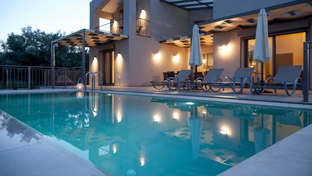 A modern poolside area with lounge chairs and umbrellas, a pool, and a building with balconies and exterior lighting. - Villa Alkistis in Lefkada (Lefkada)