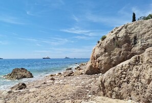 A rocky shoreline with clear blue water, boats in the distance, and a cliff with sparse vegetation. - Apartment Near Monaco Rolex Masters (Roquebrune-Cap-Martin)